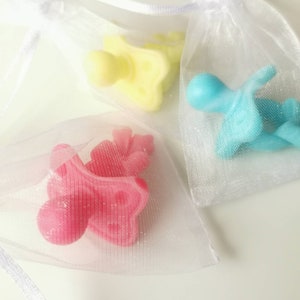 May include: Three small baby pacifier shaped silicone toys in different colors, yellow, blue, and pink, are displayed in white organza bags.