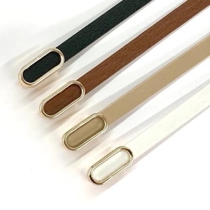 May include: Four thin belts in a row, each with a different color and a gold-colored oval buckle. The colors are dark green, brown, beige, and white. The belts are angled slightly from the top left to the bottom right.