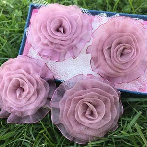 May include: Four delicate, layered, pink fabric rose flower brooches in a blue and white box. The roses are made of sheer fabric and have a soft, romantic look.