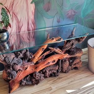 May include: A glass-top console table with a unique base made of natural driftwood and volcanic rock. The table has a rustic, organic look and would be a great addition to any home.