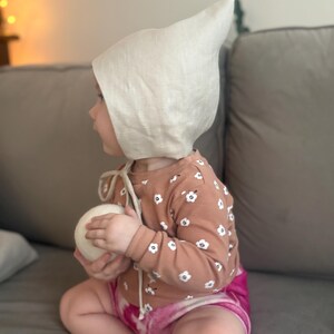 May include: A baby wearing a light beige linen bonnet with a pointed top and tie closure. The baby is also wearing a long-sleeved brown shirt with white floral patterns and pink tie-dye shorts, holding a white ball.