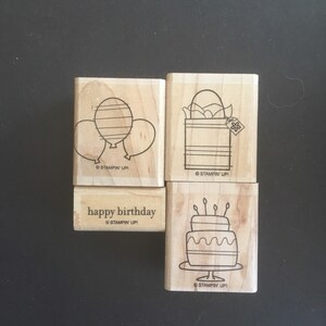May include: Four wooden stamps with black line art designs. The stamps feature a balloon design, an egg in a basket, a two-tiered cake with candles, and the words "happy birthday". The stamps are from Stampin' Up!