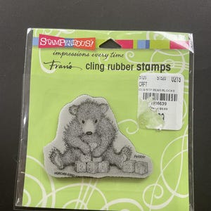 May include: A cling rubber stamp set featuring a detailed illustration of a bear cub holding blocks that spell "WE BABY". The stamp is packaged on a green backing with the text "STAMPENDOUS!" and "impressions every time".