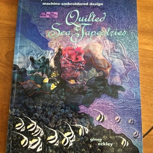 May include: Book cover with a purple, blue, and green background featuring a colorful underwater scene with fish and coral. The title is "Quilted Sea Tapestries" with the subtitle "techniques for machine-embroidered design". The author is Ginny Eckley.