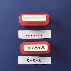 May include: Two red and white paper punch tools with a floral design. The top punch has a wavy line with hearts and the bottom punch has tulips and hearts. The punches are shown next to the paper they have punched.