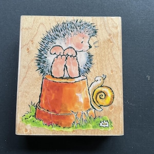 May include: A wooden stamp featuring a cartoon hedgehog sitting on a tree stump. The hedgehog has a light pink face, gray quills, and is looking down. A yellow snail is next to the stump, and green grass surrounds it.