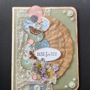 May include: A handmade greeting card with a collage design. It features layered paper circles, a butterfly, and floral accents. The text "HERE for YOU" is displayed on an oval. Embellishments include lace, pearls, and a button. The color palette includes soft pastels and earth tones.