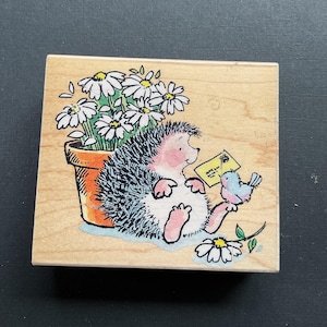 May include: A wooden stamp featuring a cartoon hedgehog with a light pink face and feet, holding a letter and sitting next to a blue bird. A pot of daisies is to the left. The stamp is on a light wood block.
