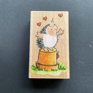 May include: A wooden stamp featuring a whimsical illustration of a hedgehog standing on a flower pot, with hearts floating above. The hedgehog has a white belly and spiky back. A ladybug is on the pot, and green grass surrounds the pot.