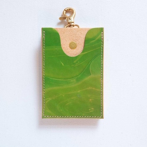 Puede incluir: Un tarjetero rectangular de cuero verde con un patrón veteado. La solapa superior es de cuero color beige y se asegura con un broche dorado. Un cierre dorado está unido en la parte superior. Los bordes están cosidos con hilo verde.