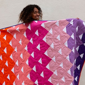 May include: A colorful quilt with a geometric pattern of white triangles and circles on a background of orange, pink, and purple. The quilt is being held up by a person.
