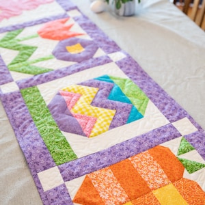 May include: A colorful patchwork table runner with a purple background and geometric designs in shades of green, yellow, orange, pink, and blue.