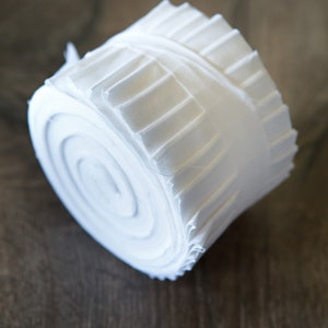 May include: A roll of white fabric, tightly wound and secured with a matching band. The fabric appears to be a soft, lightweight material, possibly cotton or linen. The roll is resting on a dark wooden surface.