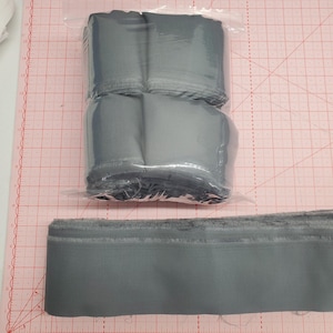 May include: A stack of four folded pieces of solid gray fabric encased in a clear plastic bag, positioned above a single long, unfolded strip of the same gray fabric. The material appears smooth with a visible selvage edge along the strip.