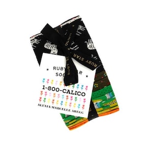 May include: A stack of fabric squares tied with a black ribbon labeled Ruby Star Society. A white tag features the text Ruby Star Society, 1-800-CALICO, and Alexia Marcelle Abegg with colorful flower icons. The fabric patterns include tigers and stripes.