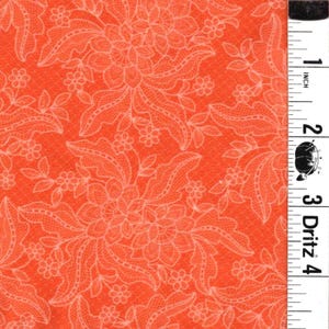 May include: A bright orange fabric featuring a repeating white lace-like floral pattern. A white measuring tape is positioned vertically along the right edge, showing a scale in inches with the Dritz brand logo clearly visible near the two-inch mark.