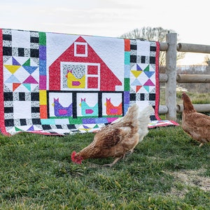May include: A colorful quilt with a red barn and chickens hanging on a wooden fence. Two brown chickens are in the grass in front of the quilt.