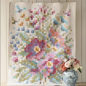May include: A large, rectangular tapestry with a pixelated floral design in shades of pink, purple, blue, and yellow. The tapestry is hanging against a light-colored wall. A vase with flowers sits in front of the tapestry.