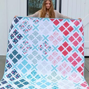 May include: A handmade quilt with a white background and a repeating pattern of diamonds and crosses. The diamonds feature floral, polka dot, and solid patterns in shades of red, blue, and teal. The quilt is held up by a person.