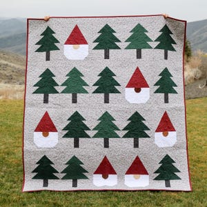 May include: A handmade quilt featuring a repeating pattern of green Christmas trees and gnome houses with red roofs and white beards. The quilt has a gray background with a subtle floral pattern and a red border. The quilt is held up outdoors.