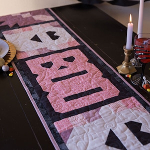 May include: A pink and black quilted table runner with the word "BOO" spelled out in black letters. The runner is on a dark wood table with two lit candles and a glass of red liquid.