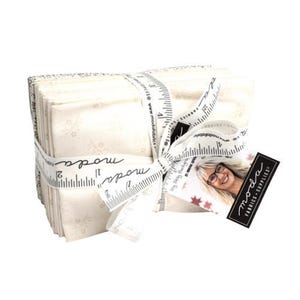 May include: A bundle of white fabric with a snowflake pattern, tied with a white ribbon and a Moda Fabrics tag. The tag has a picture of a woman with a measuring tape and the text "Moda Fabrics - Supplies".
