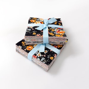 May include: Two stacks of fabric squares with a floral pattern in orange, yellow, and white on a black background. Each stack is tied with a light blue ribbon. The fabric squares are likely for quilting or other crafts.