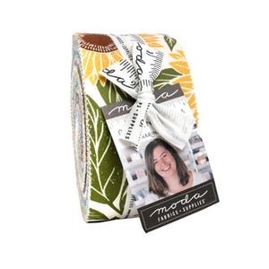 May include: A rolled fabric bundle featuring a sunflower and leaf pattern in yellow, green, and brown. The bundle is tied with a white ribbon and includes a tag with a woman's photo and the words "moda FABRICS + SUPPLIES".