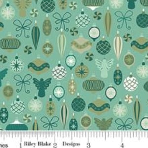 May include: A light green fabric featuring a repeating pattern of dark green, olive, and white Christmas ornaments, bows, and angel silhouettes. A white ruler at the bottom shows the scale in inches, with the text Riley Blake Designs printed.