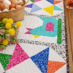 May include: A white quilted table runner with a colorful patchwork design featuring a repeating pattern of triangles. The triangles are made of different fabrics with paisley patterns in various colors, including blue, red, green, yellow, and orange. The runner is bordered with a black and white paisley trim.
