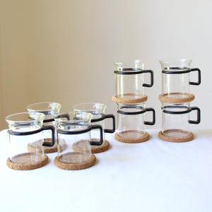 May include: A set of six clear glass mugs, each featuring a black handle and a circular black band around the upper body. Every mug rests on a round, light brown cork coaster, creating a uniform collection of drinkware for serving beverages.