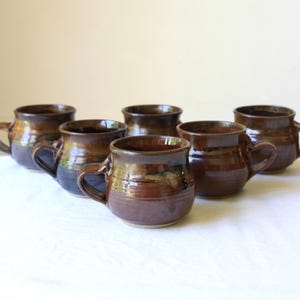 May include: A set of five rounded ceramic mugs featuring a glossy, multi-toned brown glaze with darker accents. Each mug has a sturdy handle and a wide, circular opening, showcasing a smooth, reflective surface finish across the collection.