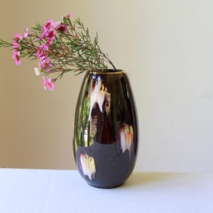 May include: A dark brown, teardrop-shaped vase with a glossy finish. The vase has white and yellow drip-like accents. Pink flowers and green stems are arranged inside the vase, creating a floral display. The vase is on a white surface.