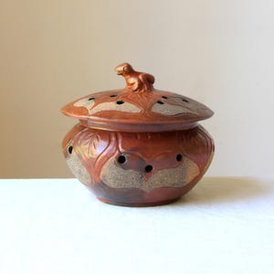 May include: A round, reddish-brown ceramic lidded jar featuring a sculpted frog figure on the lid. The body and lid are decorated with embossed leaf patterns and small, evenly spaced circular ventilation holes throughout the surface.