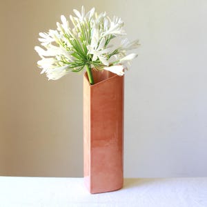 May include: A tall, rectangular vase with a unique angled opening. The vase is made of ceramic and has a light peach colour. It is filled with white flowers.