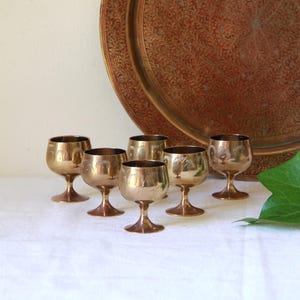 May include: Six vintage brass cordial glasses with etched designs are arranged on a white surface. A large, ornate copper-coloured tray is in the background. The glasses have a classic stemmed design.