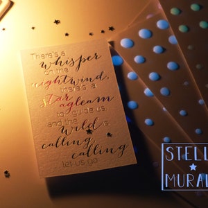 May include: A white card with gold lettering that reads "There's a whisper on the nightwind, there's a star agleam to guide us, and the wild is calling calling let us go". The card is on a brown surface with gold glitter stars scattered around it. The card is next to a stack of three sheets of paper with blue dots on them. The bottom sheet has the text "STELLA MURALS" in blue letters with a white star.