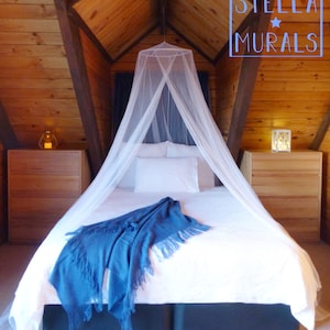 May include: A white canopy mosquito net hangs over a white bed with a blue throw blanket. The bed is in a room with wood paneling on the walls.
