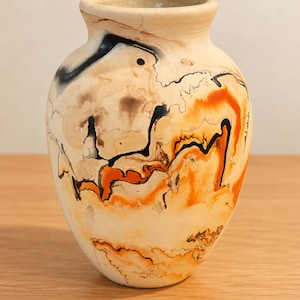 May include: A ceramic vase with a rounded body and a narrow neck, featuring a cream-colored base with swirling patterns of black and vibrant orange smoke-fired markings that create organic, flowing lines across the entire exterior surface.