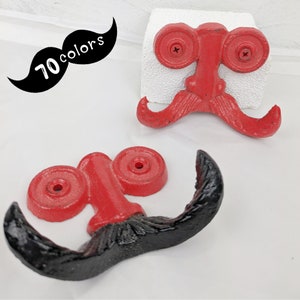 May include: Two cast iron mustache-shaped wall hooks. One hook is red with a black mustache, and the other is red. The hooks are mounted on a white surface. A black mustache graphic with the text "70 colors" is in the upper left corner.