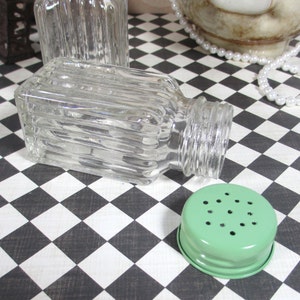 May include: Clear glass salt shaker with a ribbed design, lying on its side. A light green metal lid with small holes is next to the shaker. The background is a black and white checkered pattern.