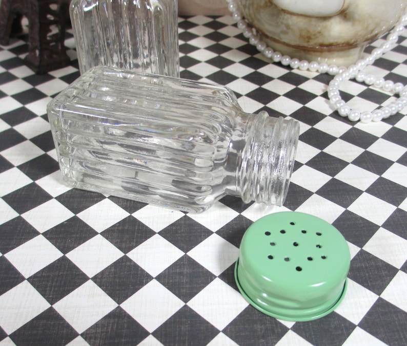 May include: Clear glass salt shaker with a ribbed design, lying on its side. A light green metal lid with small holes is next to the shaker. The background is a black and white checkered pattern.