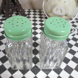 May include: A pair of vintage glass salt and pepper shakers. Each shaker has a clear, ribbed glass body and a light green metal lid with small holes. The shakers are square-shaped with a twisted design.