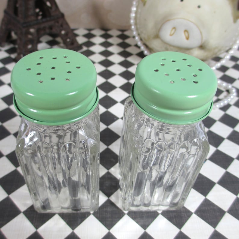 May include: A pair of vintage glass salt and pepper shakers. Each shaker has a clear, ribbed glass body and a light green metal lid with small holes. The shakers are square-shaped with a twisted design.