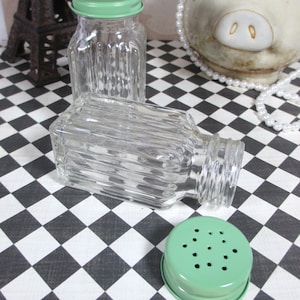 May include: Clear glass salt and pepper shakers with ribbed sides and light green metal lids. One shaker is upright, another is on its side, and a lid is separate. The background features a black and white checkered pattern.