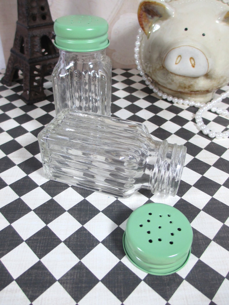 May include: Clear glass salt and pepper shakers with ribbed sides and light green metal lids. One shaker is upright, another is on its side, and a lid is separate. The background features a black and white checkered pattern.