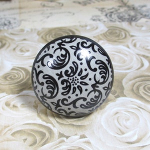 May include: A decorative, round cabinet knob with a silver-gray base and a black, ornate floral pattern. The knob is a sphere shape and is designed for use on drawers or cabinets. The design is symmetrical and detailed.