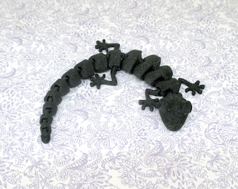 May include: A black articulated lizard figurine with a textured surface. The lizard features a segmented body design that allows for flexible movement, a wide head, and four splayed feet with distinct toes, all rendered in a matte black finish.