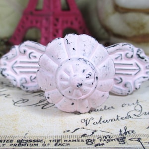 May include: Three distressed, pink cabinet knobs with black accents. The center knob is round with a floral design, while the flanking knobs are rectangular with arrow details. Decorative hardware for furniture.
