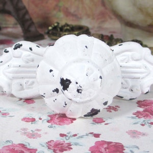 May include: Three white, distressed-finish cabinet knobs with a floral design. The knobs have a round, fluted center and decorative edges. The white paint is chipped, revealing the dark base material. The knobs are arranged on a floral patterned surface.